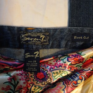 May include: Dark blue denim jeans with a boot cut style. The interior label reads Seven7, size 29, 98% cotton, 2% spandex, made in Hong Kong. A colorful satin fabric with a floral paisley pattern in shades of pink, red, blue, and yellow is tucked inside.