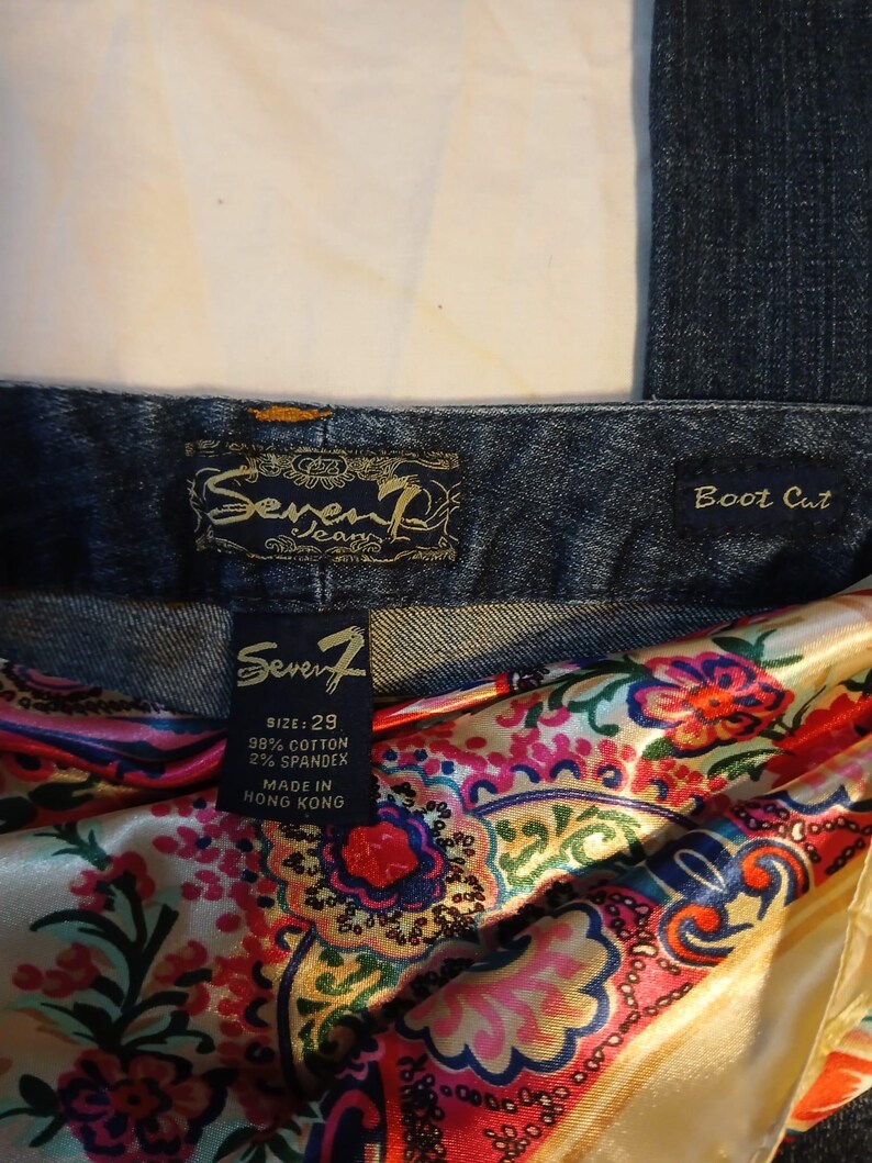 May include: Dark blue denim jeans with a boot cut style. The interior label reads Seven7, size 29, 98% cotton, 2% spandex, made in Hong Kong. A colorful satin fabric with a floral paisley pattern in shades of pink, red, blue, and yellow is tucked inside.
