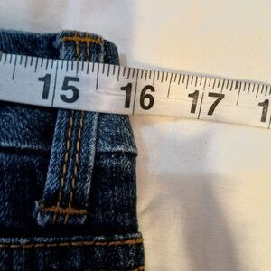 May include: A pair of blue denim jeans featuring a waistband with belt loops and orange contrast stitching. A white measuring tape is positioned across the top of the waistband, indicating a measurement of approximately 16 inches across.