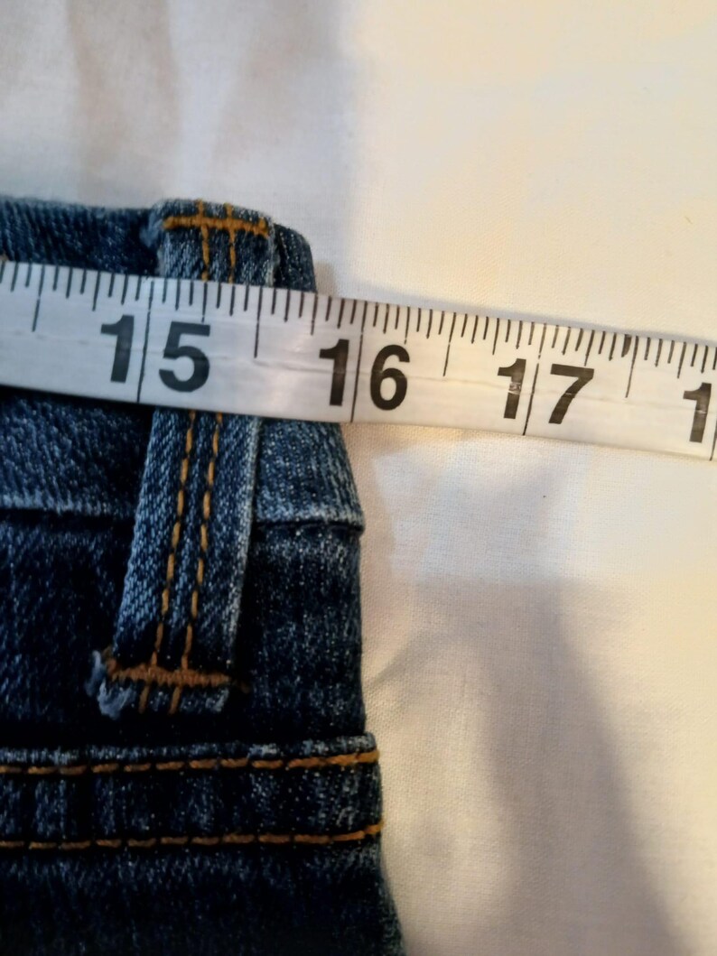 May include: A pair of blue denim jeans featuring a waistband with belt loops and orange contrast stitching. A white measuring tape is positioned across the top of the waistband, indicating a measurement of approximately 16 inches across.