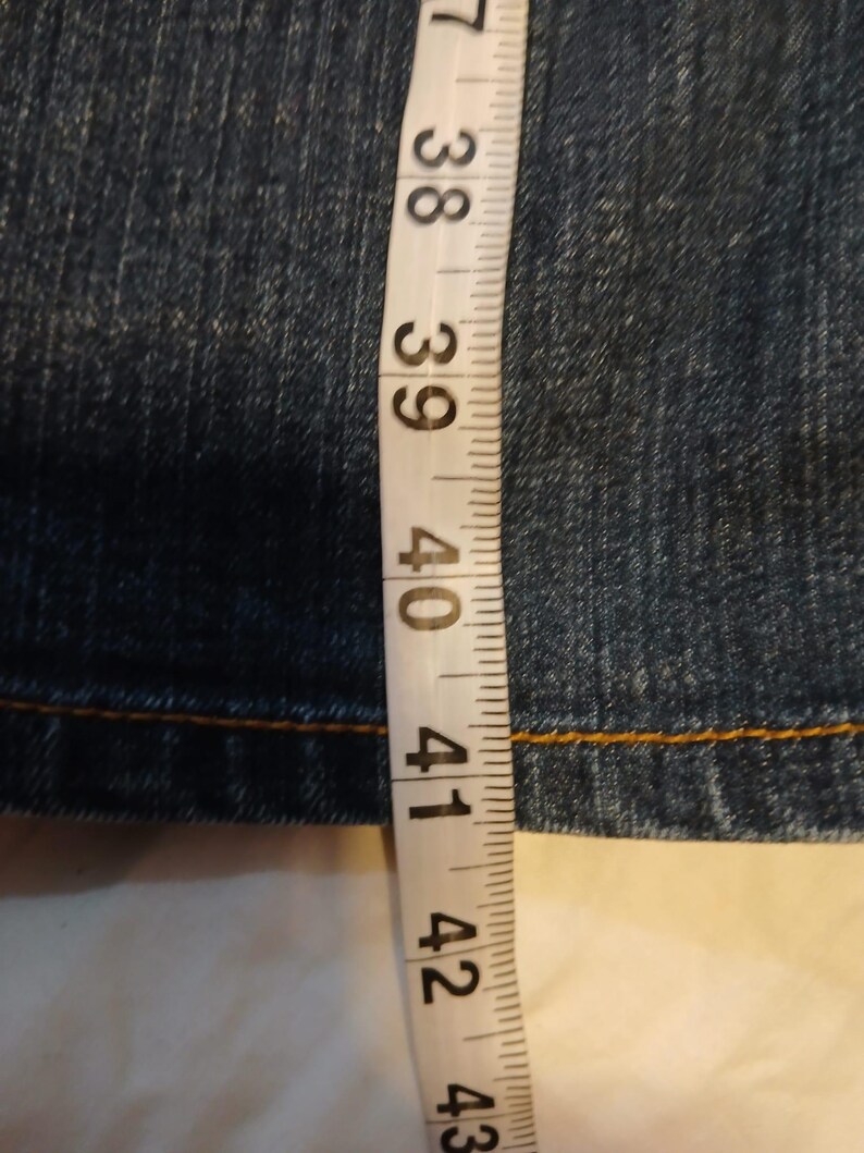 May include: A dark blue denim fabric with visible stitching, positioned next to a white measuring tape that indicates a length measurement extending past 40 inches, with the tape showing increments from 37 to 43 inches clearly displayed.