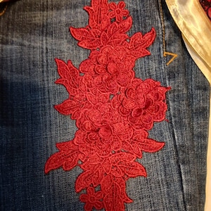 May include: A pair of blue denim jeans featuring a large, intricate red floral embroidery patch on the front. The embroidery consists of detailed flowers and leaves. A colorful patterned fabric with pink, orange, and gold tones is visible.