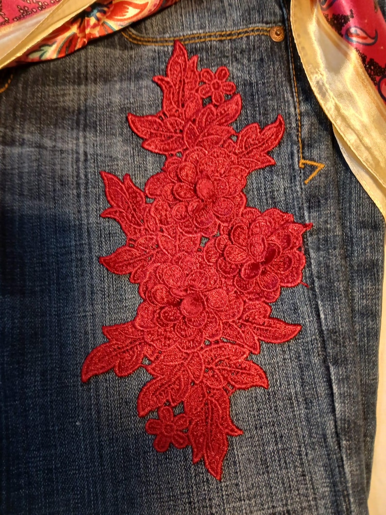 May include: A pair of blue denim jeans featuring a large, intricate red floral embroidery patch on the front. The embroidery consists of detailed flowers and leaves. A colorful patterned fabric with pink, orange, and gold tones is visible.
