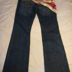 May include: Dark blue denim jeans featuring a bootcut leg silhouette and a back pocket with a stitched T-shaped logo. A colorful silk-like scarf with floral and paisley patterns in red, pink, blue, and gold is draped across the waistband.