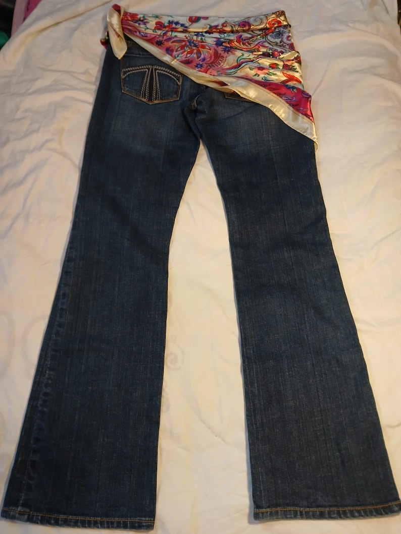 May include: Dark blue denim jeans featuring a bootcut leg silhouette and a back pocket with a stitched T-shaped logo. A colorful silk-like scarf with floral and paisley patterns in red, pink, blue, and gold is draped across the waistband.