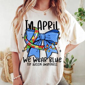 May include: Cream-colored t-shirt with the text "In April We Wear Blue for autism awareness." The design features a blue bow with a puzzle ribbon and heart accents, promoting autism awareness.