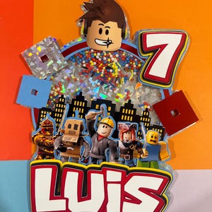 May include: A colorful cake topper featuring Roblox characters, the number 7, and the name "LUIS". The topper has a cityscape silhouette and includes a clear section filled with colorful sprinkles and confetti. The design is set against a vibrant orange background.