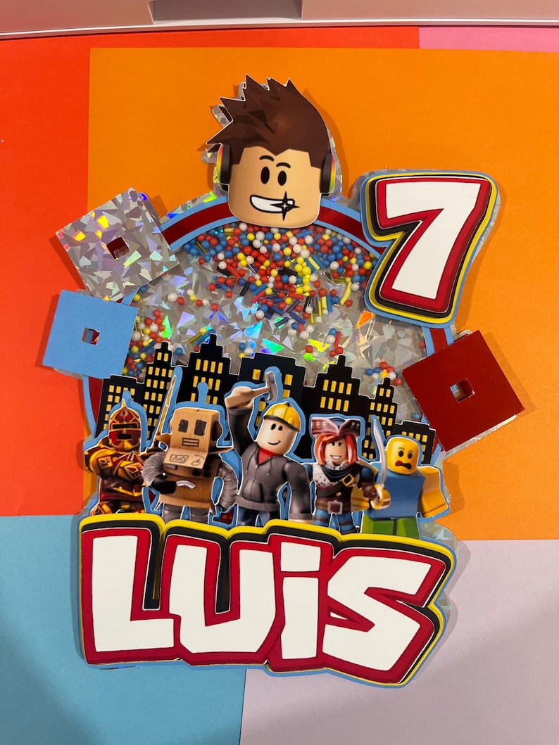 May include: A colorful cake topper featuring Roblox characters, the number 7, and the name "LUIS". The topper has a cityscape silhouette and includes a clear section filled with colorful sprinkles and confetti. The design is set against a vibrant orange background.