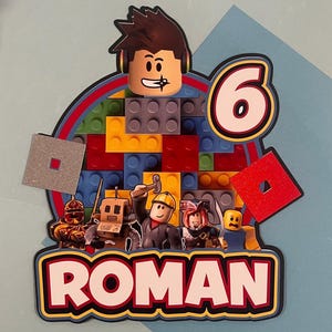 May include: A colorful birthday cake topper featuring a Roblox character design. The topper includes a large number 6, the name "ROMAN", and various Roblox figures. The background is made of colorful building blocks.