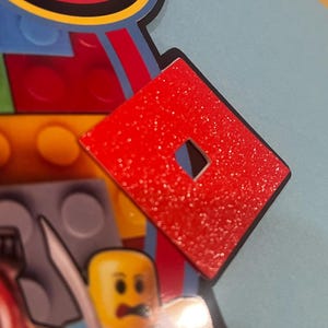 May include: A red, glittery Roblox logo with a square cut-out in the center. The logo is set against a backdrop of colorful Lego-themed elements, including a Lego figure and building blocks. The image has a close-up perspective.