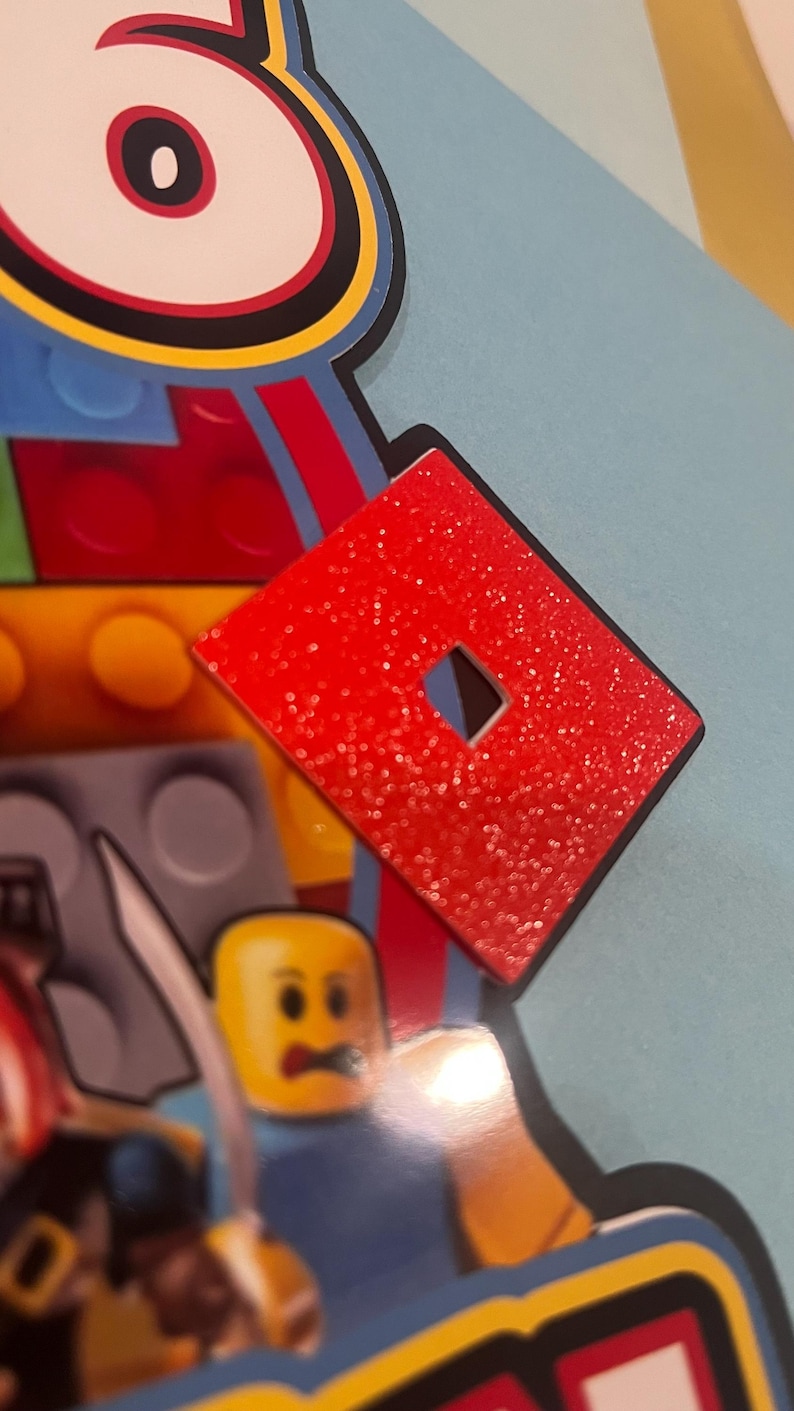 May include: A red, glittery Roblox logo with a square cut-out in the center. The logo is set against a backdrop of colorful Lego-themed elements, including a Lego figure and building blocks. The image has a close-up perspective.