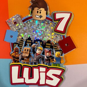 May include: A colorful birthday cake topper featuring a Roblox character head, the number 7, and the name "LUIS". The design includes various Roblox avatars and city skyline silhouettes, with a background of sparkling confetti.