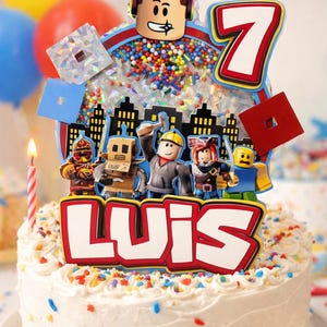 May include: A white frosted cake with colorful sprinkles, topped with a personalized Roblox-themed cake topper. The topper features the name "LUIS", the number "7", and various Roblox character figures. A lit candle and balloons are in the background.