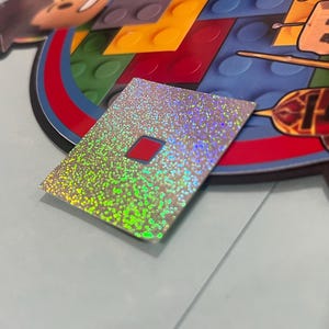 May include: A holographic sticker with a red square in the center. The sticker has a silver and green iridescent finish. The sticker is rectangular and is placed on a light blue surface. The sticker is next to a colorful graphic.
