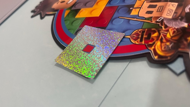 May include: A holographic sticker with a red square in the center. The sticker has a silver and green iridescent finish. The sticker is rectangular and is placed on a light blue surface. The sticker is next to a colorful graphic.