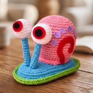 May include: A handmade crochet snail figurine with pink, blue, red, and white details. The snail has large, expressive eyes and a shell with purple spots. The base is blue with a green border, and the snail is sitting on a wooden surface.