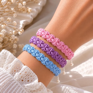 Può includere: Quattro braccialetti colorati intrecciati, blu, viola e rosa, sono esposti su un polso. I braccialetti hanno un design strutturato e annodato. Lo sfondo è morbido e chiaro, con accenti floreali.