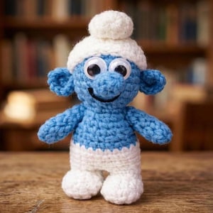 May include: A handmade crochet Smurf doll, featuring a blue body, white pants, and a white hat with a pom-pom. The doll has large googly eyes and a stitched black smile. The background is blurred, suggesting a wooden surface.