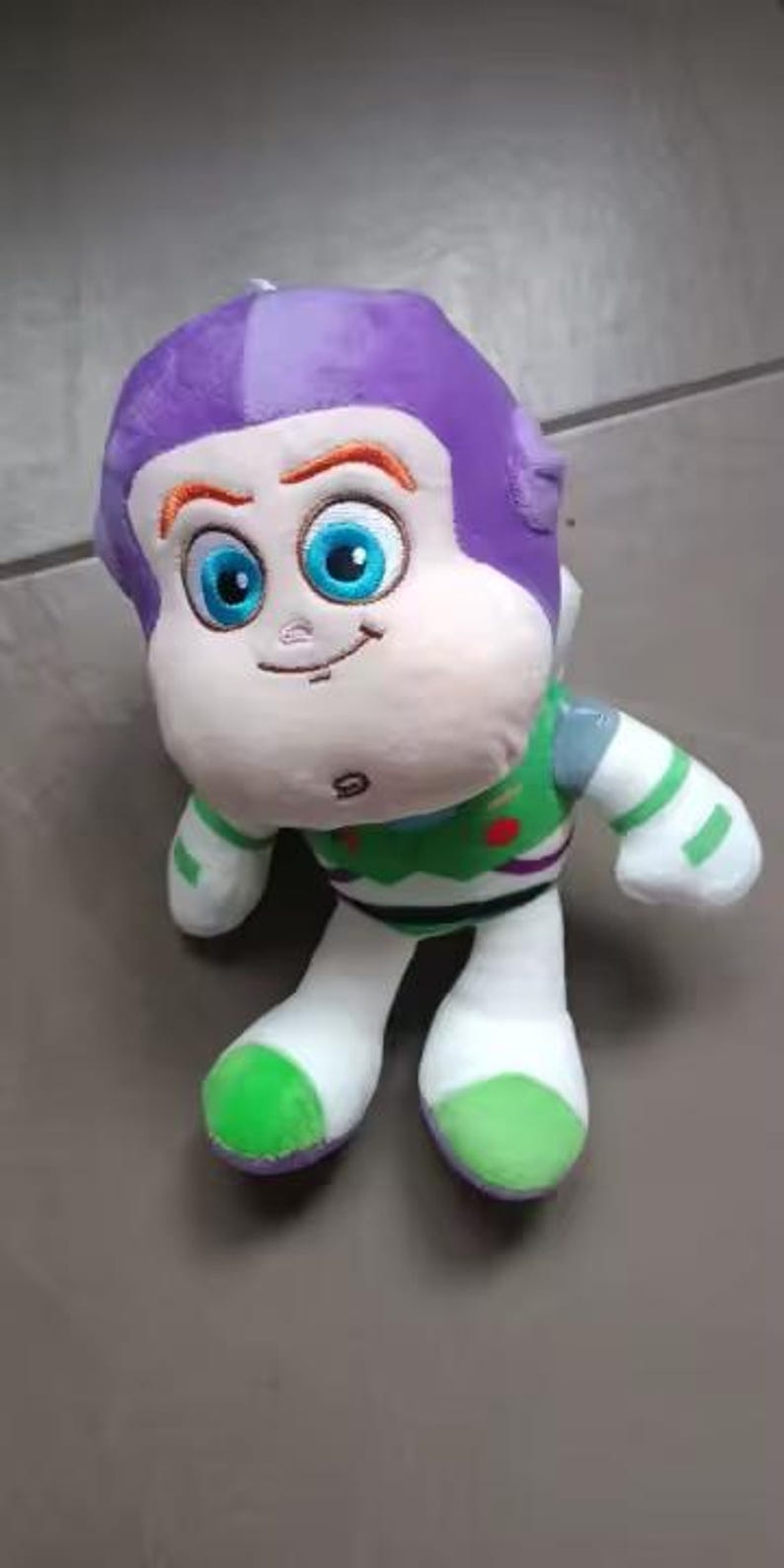 May include: A plush Buzz Lightyear doll, with a purple helmet, white face, and blue eyes. The doll wears a green and white spacesuit with red and green accents. The toy is designed to resemble the character from the animated film.