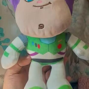 May include: A soft Buzz Lightyear doll with a purple head, tan face, and large blue eyes. The doll is dressed in a green and white spacesuit with purple and green details. The toy is designed for children and is soft to the touch.