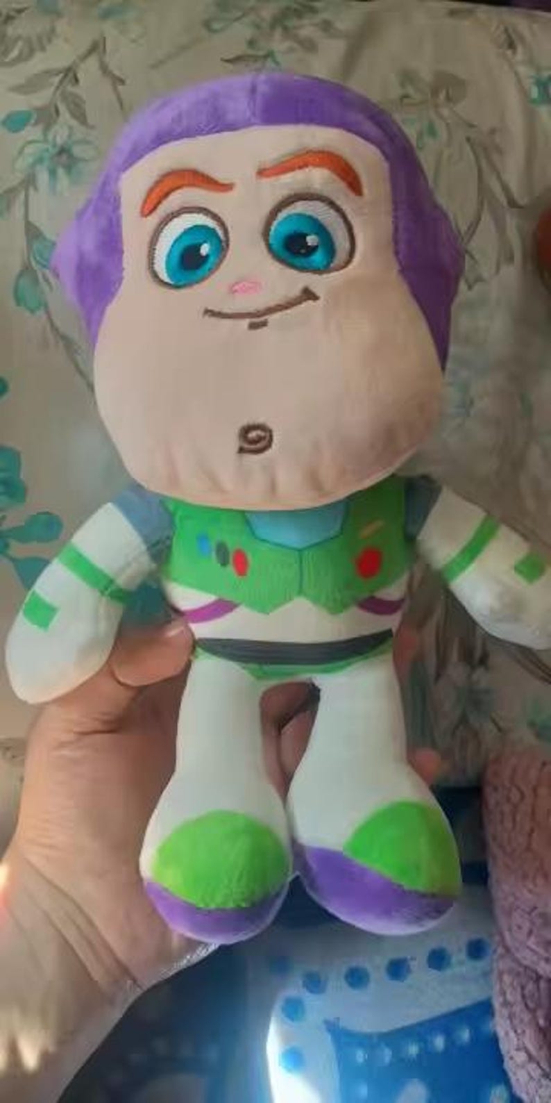 May include: A soft Buzz Lightyear doll with a purple head, tan face, and large blue eyes. The doll is dressed in a green and white spacesuit with purple and green details. The toy is designed for children and is soft to the touch.