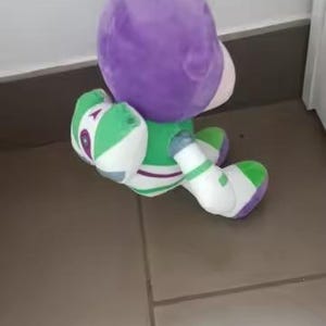 May include: A soft toy of the character Buzz Lightyear from the film Toy Story. The toy features a purple helmet, a green and white spacesuit, and white boots. The toy is being held by a hand.