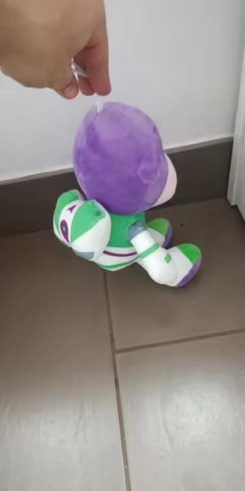 May include: A soft toy of the character Buzz Lightyear from the film Toy Story. The toy features a purple helmet, a green and white spacesuit, and white boots. The toy is being held by a hand.