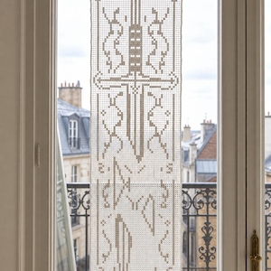 May include: A rectangular off-white crochet panel featuring a central sword motif rendered in a grid-like pattern. The design includes geometric borders and symmetrical abstract shapes surrounding the sword, suspended by a thin string.