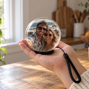 May include: A clear spherical glass ornament featuring a printed photograph of a smiling couple wearing sunglasses at the beach. The sphere has a black base and a black wrist strap attached to the side, allowing it to be held in the palm of a hand.