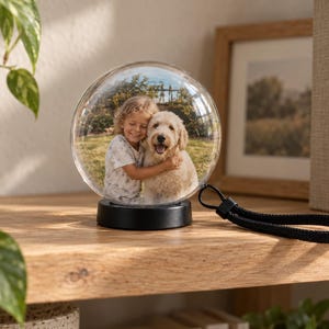 May include: A clear spherical glass ornament featuring a photographic print of a child hugging a golden-colored dog. The sphere is mounted on a circular black base and includes a black braided wrist strap attached to the side of the base.