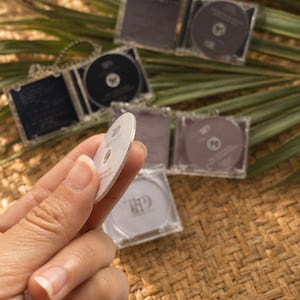 May include: A small, circular, white disc featuring a dark logo and text, held between fingers. Several clear, square plastic cases containing similar circular discs with dark labels are arranged in the background, displaying the same logo and text.