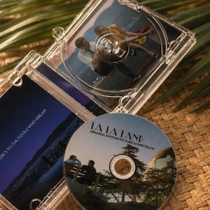 May include: A clear plastic CD case for the La La Land original motion picture soundtrack. The disc features an image of two people on a bench near an observatory. The case insert displays the text Here's to the fools who dream over a night sky.