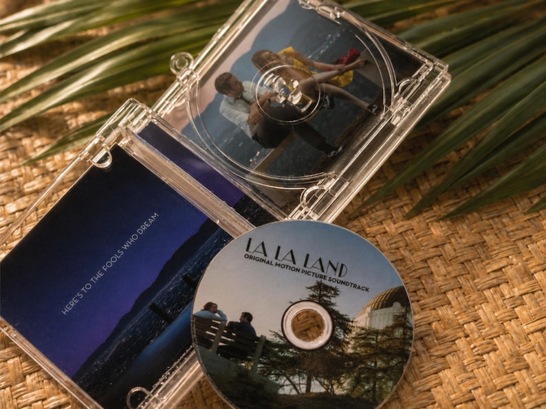 May include: A clear plastic CD case for the La La Land original motion picture soundtrack. The disc features an image of two people on a bench near an observatory. The case insert displays the text Here's to the fools who dream over a night sky.