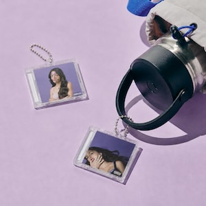May include: Two square clear plastic keychains featuring miniature album cover art of a person with long dark hair against a purple background, attached to a silver ball chain, alongside a stainless steel water bottle with a black lid and handle.