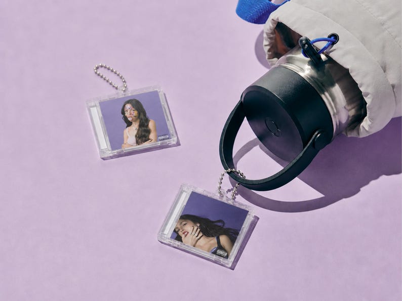 May include: Two square clear plastic keychains featuring miniature album cover art of a person with long dark hair against a purple background, attached to a silver ball chain, alongside a stainless steel water bottle with a black lid and handle.
