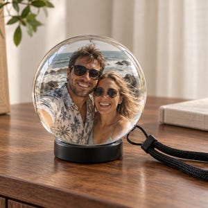 May include: A spherical glass ornament featuring a photograph of a smiling couple wearing sunglasses on a beach. The clear globe is mounted on a solid black circular base and includes a black braided wrist strap attached to the side of the base.