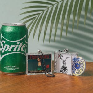 May include: A green Sprite soda can next to two miniature clear plastic cases featuring NewJeans album art. One case has a black ball chain, and the other includes a small circular disc with the text "SWEET" printed in blue and yellow on the front.