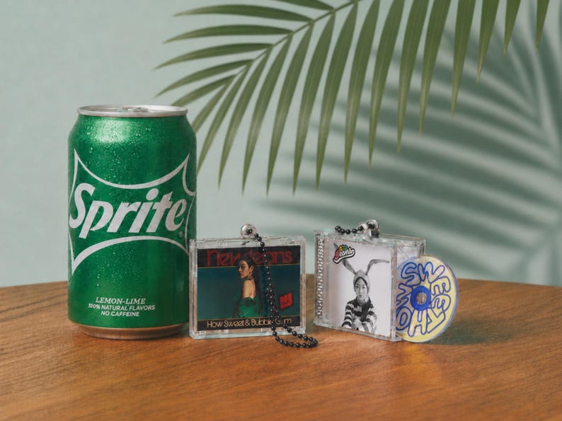 May include: A green Sprite soda can next to two miniature clear plastic cases featuring NewJeans album art. One case has a black ball chain, and the other includes a small circular disc with the text "SWEET" printed in blue and yellow on the front.