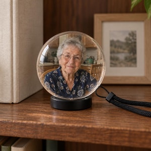 May include: A clear glass sphere ornament featuring a photographic portrait of an elderly person with gray hair and glasses wearing a dark blue floral shirt. The sphere is mounted on a circular black base and includes a black braided wrist strap.