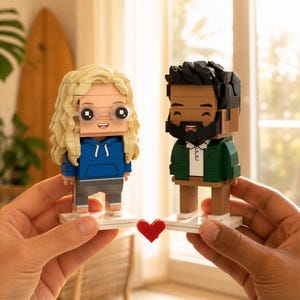 May include: Two custom brick-built figurines standing on white square bases. The left figure has blonde hair, glasses, and a blue hoodie with gray pants. The right figure has dark hair, a beard, a green jacket, and a white shirt. A small red heart sits between them.