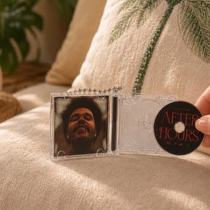 May include: A miniature clear plastic CD case keychain featuring a portrait of The Weeknd on the cover and a black disc inside with the text After Hours printed in red. The keychain includes a silver ball chain attached to the top hinge.