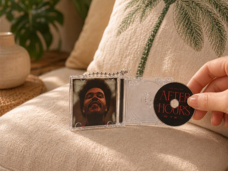 May include: A miniature clear plastic CD case keychain featuring a portrait of The Weeknd on the cover and a black disc inside with the text After Hours printed in red. The keychain includes a silver ball chain attached to the top hinge.