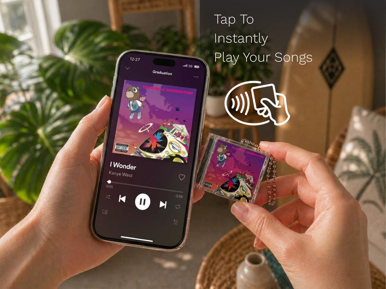 May include: A smartphone displaying a music streaming interface with the album art for Graduation by Kanye West, alongside a small, square, clear acrylic keychain featuring the same album artwork and a metal ball chain, with text reading Tap To Instantly Play Your Songs.
