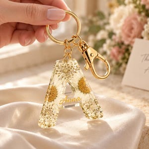 May include: A clear resin keyring in the shape of the letter A, embedded with small white daisy flowers and gold glitter. The keyring features a gold-toned metal split ring and a lobster clasp, with the name Anna printed in gold script.