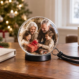 May include: A circular glass ornament featuring a printed photograph of a family of four opening a red gift box. The ornament is mounted on a black circular base and includes a black braided cord loop for hanging, creating a decorative display piece.