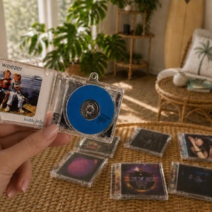 May include: A miniature clear plastic CD case keychain featuring the Weezer Buddy Holly album cover art on the front and a solid blue disc inside. The keychain is held open to display the album artwork and the disc, with several other similar miniature CD case keychains arranged on a surface below.