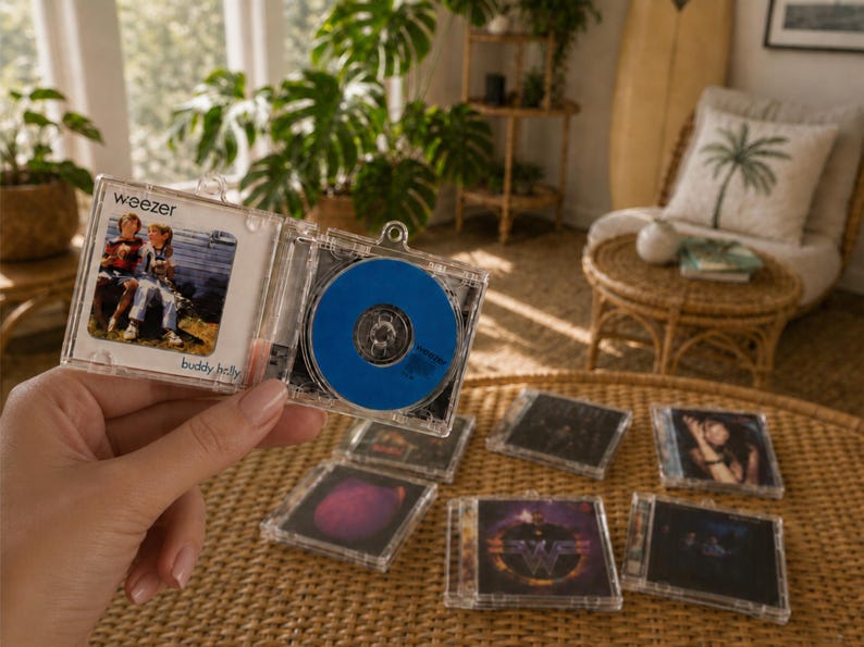May include: A miniature clear plastic CD case keychain featuring the Weezer Buddy Holly album cover art on the front and a solid blue disc inside. The keychain is held open to display the album artwork and the disc, with several other similar miniature CD case keychains arranged on a surface below.