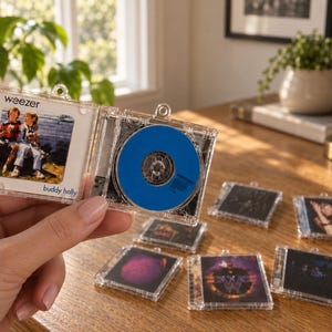 May include: A miniature clear plastic CD case ornament featuring the Weezer Buddy Holly album cover art on the front and a bright blue disc inside. The case has a small loop at the top for hanging. Several other similar miniature CD ornaments are visible.