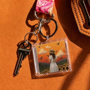 May include: A square clear acrylic keychain featuring an illustration of a person in a white dress and straw hat standing in a sunflower field under a sunset sky with birds, attached to a silver metal keyring and a pink patterned fabric strap.