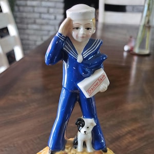 May include: A ceramic Cracker Jack figurine featuring a sailor saluting, holding a box labeled "Cracker Jack." A small dog stands nearby. The base is popcorn and almonds, with "Cracker Jack" around the rim.